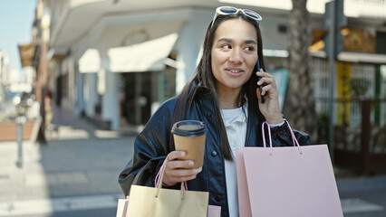 Young beautiful hispanic woman talking on smartphone holding shopping bags and coffee at coffee shop terrace