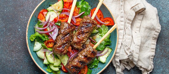 Grilled skewer meat beef kebabs on sticks served with fresh vegetables salad on plate on rustic concrete background from above. Traditional Middle Eastern and Turkish dish Kebab