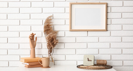 Workplace with wooden hand, books, cup, pampas grass in vase and clock near white brick wall with hanging blank photo frame