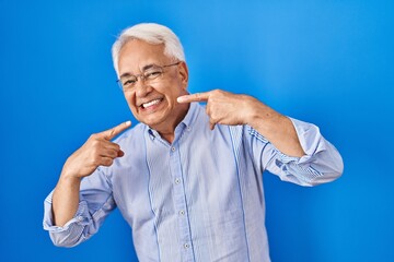 Hispanic senior man wearing glasses smiling cheerful showing and pointing with fingers teeth and mouth. dental health concept.