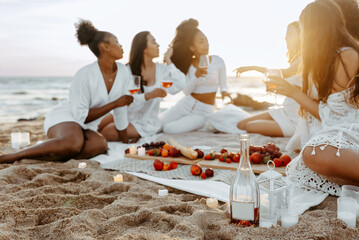 Young beautiful women having summer picnic with wine and fruits on the beach at sunset, ladies...