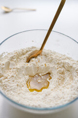 Close-up of beautiful ingredients. Glass bowl with white flour and liquid inside, with a spoon for mixing the dough on a light background. The process of cooking at home