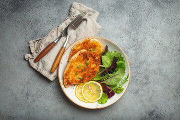 Crispy panko breaded fried chicken fillet with green salad and lemon on plate with cutlery on gray...