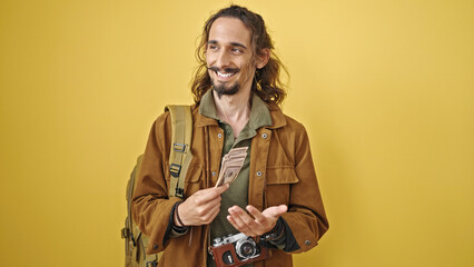 Young hispanic man tourist wearing backpack counting dollars over isolated yellow background