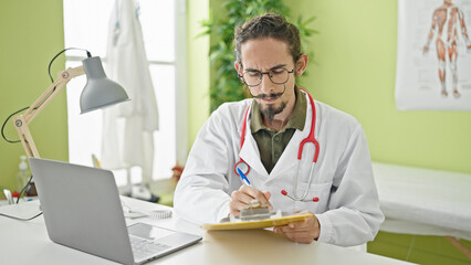 Young hispanic man doctor using laptop writing on clipboard at clinic