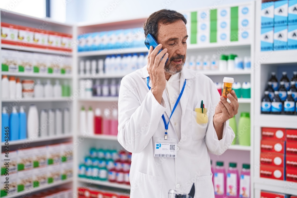 Sticker Middle age man pharmacist holding pills bottle talking on smartphone at pharmacy