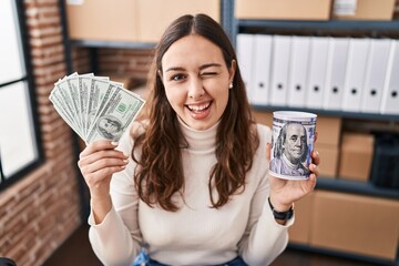 Young hispanic woman working at small business ecommerce holding money and piggy bank winking...