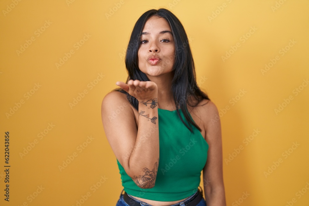 Sticker brunette woman standing over yellow background looking at the camera blowing a kiss with hand on air