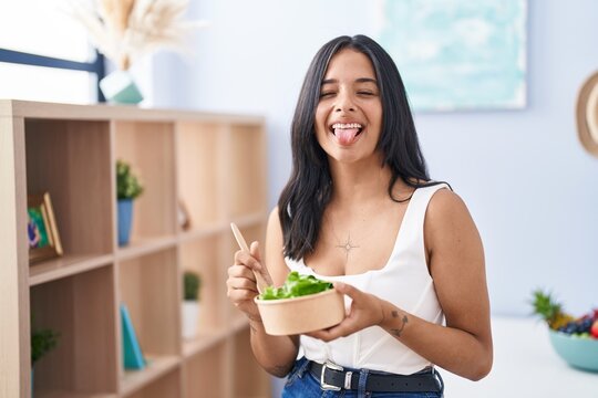 Brunette woman eating a salad at home sticking tongue out happy with funny expression.