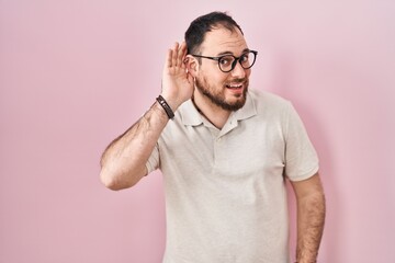 Plus size hispanic man with beard standing over pink background smiling with hand over ear listening an hearing to rumor or gossip. deafness concept.