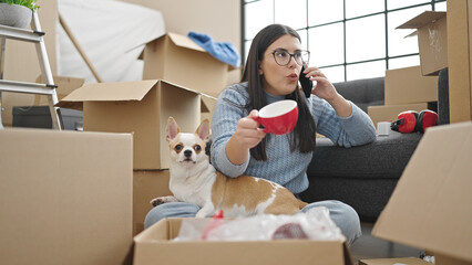 Young hispanic woman with chihuahua dog unpacking cardboard box speaking on the phone complaining...