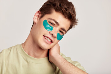 Young handsome smiling guy with brown hair applying under eye hydrogel patches. Caucasian millennial man practices skin care routine to keep healthy and youthful looking. Studio portrait.