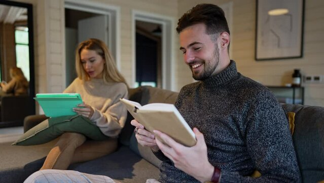 Laughing positive man reading book out loud as woman scrolling digital tablet touchscreen sitting on sofa at background. Happy Caucasian boyfriend enjoying hobby resting with girlfriend indoors