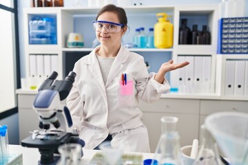 Hispanic girl with down syndrome working at scientist laboratory smiling cheerful presenting and pointing with palm of hand looking at the camera.