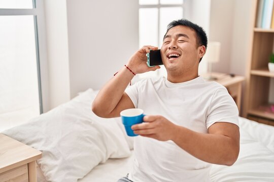 Young chinese man talking on the smartphone drinking coffee sitting on bed at bedroom