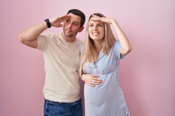 Young couple expecting a baby standing over pink background very happy and smiling looking far away with hand over head. searching concept.