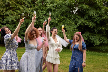 Obraz premium Happy charming bride in white dress with white veil and bridesmaids with pink veils raise champagne glasses and laugh merrily. Cheerful bachelorette party in the summer park.