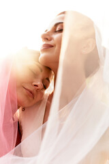 A happy charming bride in a white dress with a white veil and a bridesmaid with a pink veil hugging...