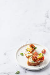 Bruschetta sandwiches with tomatoes, cream cheese, olive oil and basil on a plate on white marble background, copy space. Traditional italian antipasti