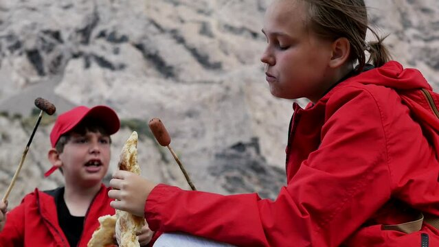 Portrait of cheerful teenagers in red clothes on a picnic in the mountains frying sausages on fire near the fire and eating fresh bread puri. Active teenagers travel and love adventure
