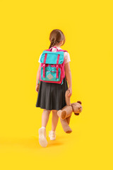 Little schoolgirl with backpack and toy bear on yellow background, back view