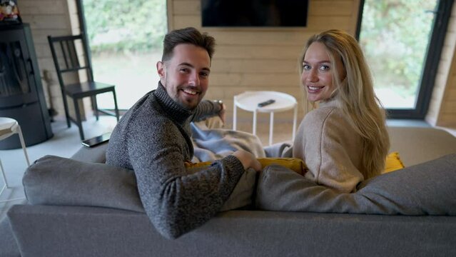 Shooting over shoulder of happy beautiful couple sitting on sofa talking turning looking at camera smiling. Loving Caucasian handsome man and charming woman posing on couch in living room