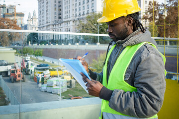 young black engineer man standing supervising the construction site making notes. - obrazy, fototapety, plakaty