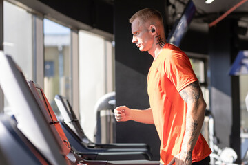 young athlete man goes in for sports on treadmill in fitness club, listening music in wireless headphones. European guy on workout in gym