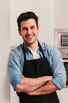 Vertical portrait of handsome caucasian man wearing an apron standing in the kitchen, smiling and crossing arms looking at camera. Front view of happy handsome male leaning against a wall at home