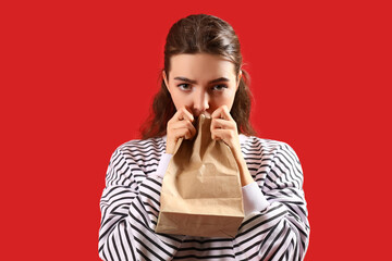 Young woman with paper bag having panic attack on red background