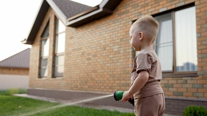 Funny little boy is watering lawn plants in the backyard of a garden house. Boy with hose. A little...