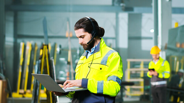 Caucasian male young engineer in goggles and yellow uniform standing in factory and talking on mobile phone. Engineering concept. Inside. Man speaking on cellphone with folder of documents at plant.
