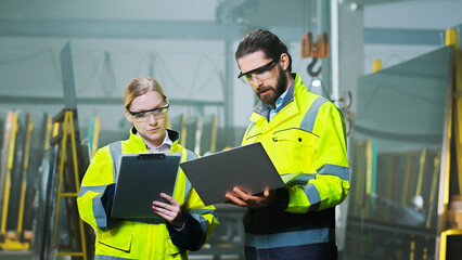 Caucasian man and woman colleagues at manufactory talking and discussing work with laptop computer and documents or plan. Production plant. Indoor. Male and female co-workers at factory.