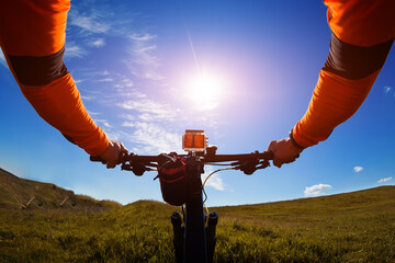 Naklejka na ściany i meble Hands in orange jacket holding handlebar of a bicycle with green meadow on background