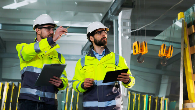 Caucasian men colleagues talking and planning work. Males walking at factory. Work with folder of documents and tablet device at manufactory. Production co-working. Discussion of technicians. Indoor.