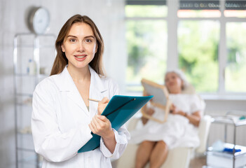 Smiling female doctor holding folder with documents and looking at camera behind woman patient on couch in clinic.