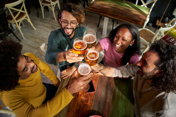 Group of multirracial young people toasting with a pint of beer and celebrating happy hours in a bar indoors. Friends drink alcohol together having fun and smiling. Cheers of gathering latin students
