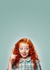 Small ginger red curly hair girl with freckles. She looks cute and innocent, but is probably little rascal. Making funny face, pointing finger to empty copy space above, vertical banner. Generative AI