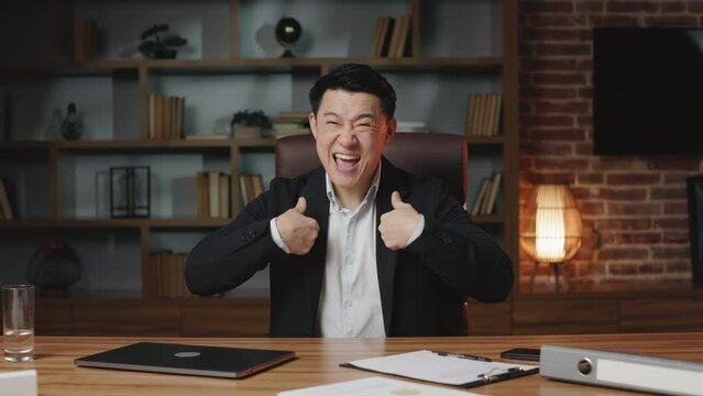 Excited asian entrepreneur dressed in white shirt and formal black suit bragging business success while sitting in rolling office chair. Proud man raising thumbs up and pointing fingers at himself.