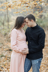 Fototapeta premium portrait of a happy young family, husband and pregnant wife, standing against the background of beautiful light in the autumn forest, looking at each other