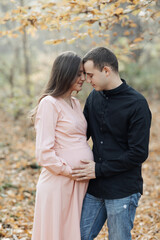 portrait of a happy young family, husband and pregnant wife, standing against the background of beautiful light in the autumn forest, looking at each other