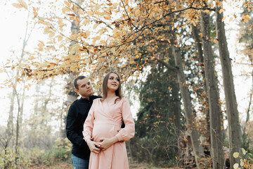 Fototapeta premium A man and his pregnant wife embrace in the forest at sunset, waiting for the birth of a child. Happy family concept.