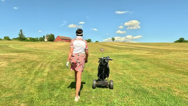 Mature woman playing golf. Female golfer walking on golf course with automatic electric golf cart. Beautiful sunny landscape, with green hills and blue sky.