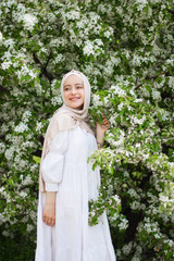 A young Muslim woman in a white dress in front of a flowering tree. Medium plan. Blooming apple tree. Pretty modest Muslim woman. European Muslim. Tenderness. Beauty.