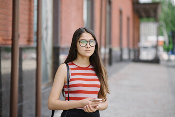 Fototapeta premium Young Asian woman with glasses hold smartphone in hands. Young woman on city background.