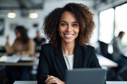 Generative AI image of portrait of smiling young African American female employee looking at camera while sitting at table with laptop and working in blurred office with colleagues