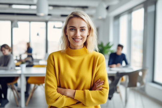 Generative AI image of portrait of smiling young female employee in sweater looking at camera while standing with hands crossed in blurred and illuminated office with colleagues