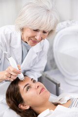 Obraz na płótnie Canvas Mature female dermatologist performing cosmetology procedure for young woman, RF lifting and vacuum massage for facial skin therapy in clinic