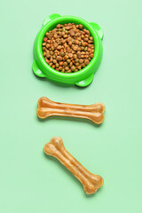Bowl with dry dog food and chew bones on color background