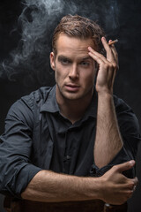 Attractive guy in dark shirt sits on a chair on the black background in the studio. He holds a cigarette in left hand and looks in front of himself. Smoke swirls around man. Vertical low-key photo.